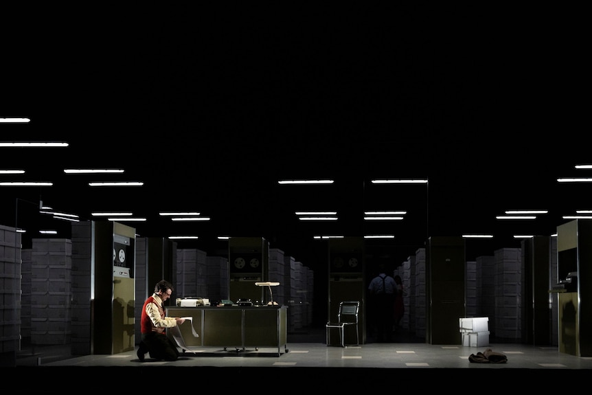 On stage, Dan Spielman, 47, in glasses, kneels on the ground in front of a desk and chair, looking over pages of code.