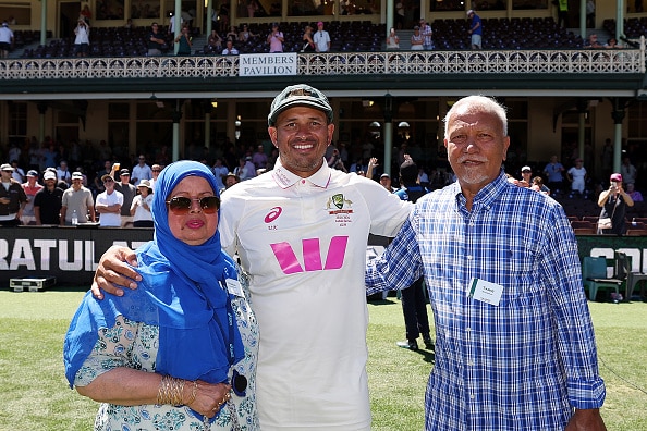 Usman Khawaja stands in crickets whites with his arms around mother and father. A crowd behind fills the stadium stands.