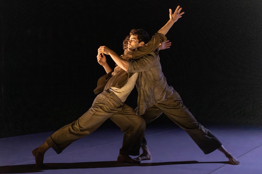 On stage, two dancers lunge into each other, knees bent, their upper torsos touching, arms in the air.