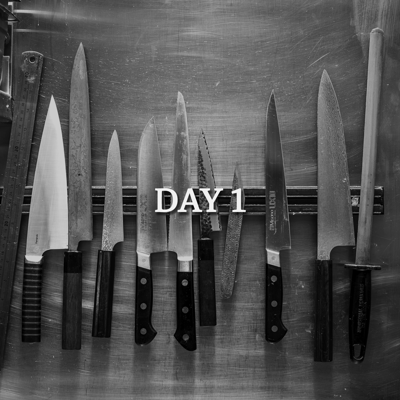 Black and white photo of various kitchen knives attached to magnetic knife rack on wall in commercial kitchen.