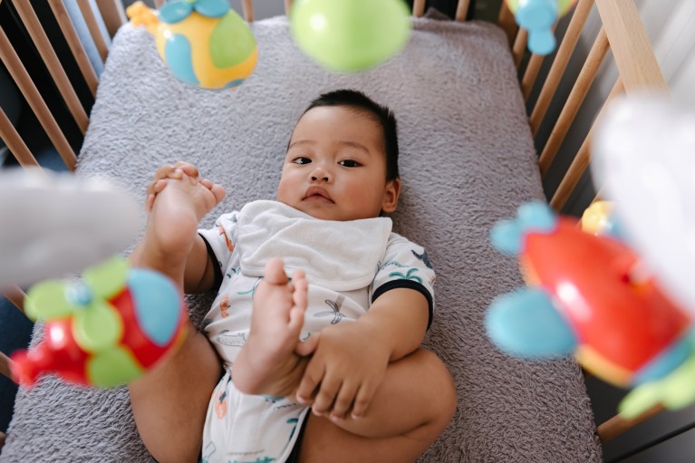 Close-up of a baby lying in a crib, holding both feet up, with colourful hanging toys blurred in the foreground