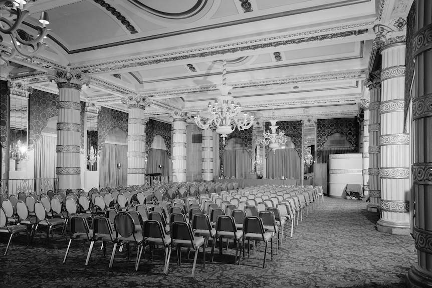 Black and white interior of a hotel conference room, chairs lined up under two chandeliers.