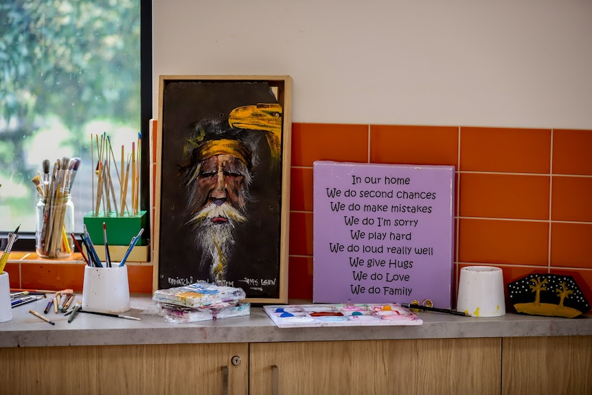 Art supplies, a sign and a painting of an older Indigenous man's face inside an art room with a window to an outside garden