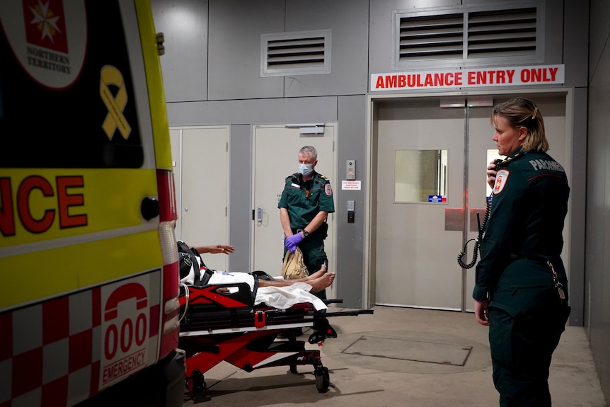 The back corner of an ambulance with a stretcher behind it and legs of a patient visible and paramedics standing nearby