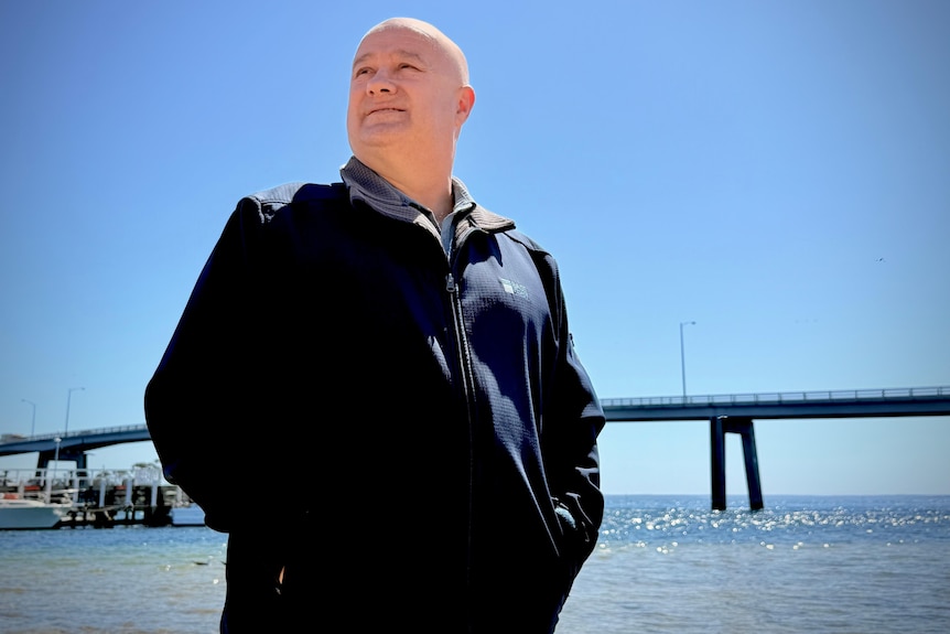 Image of a man in a black jacket looking off into the distance, standing before a blue sky.