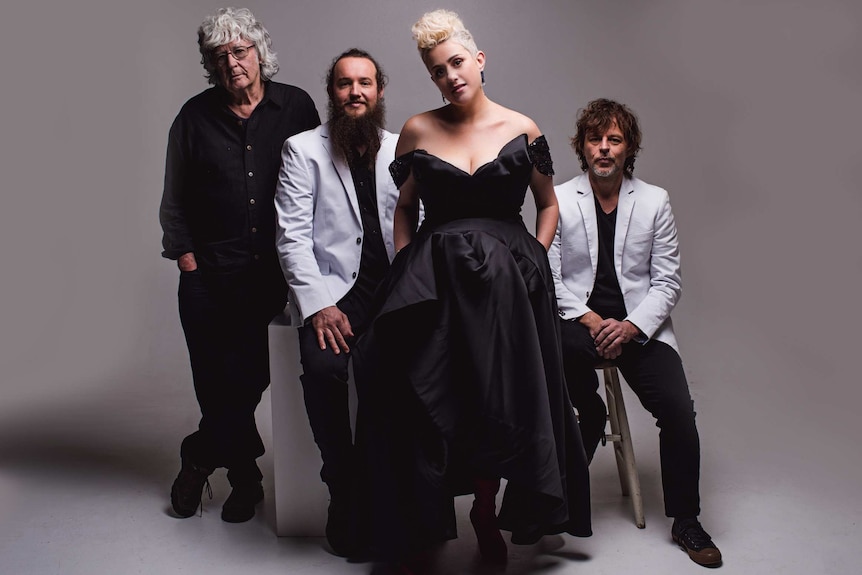 A woman and three men in formal clothing pose for a photo on a blank backdrop.