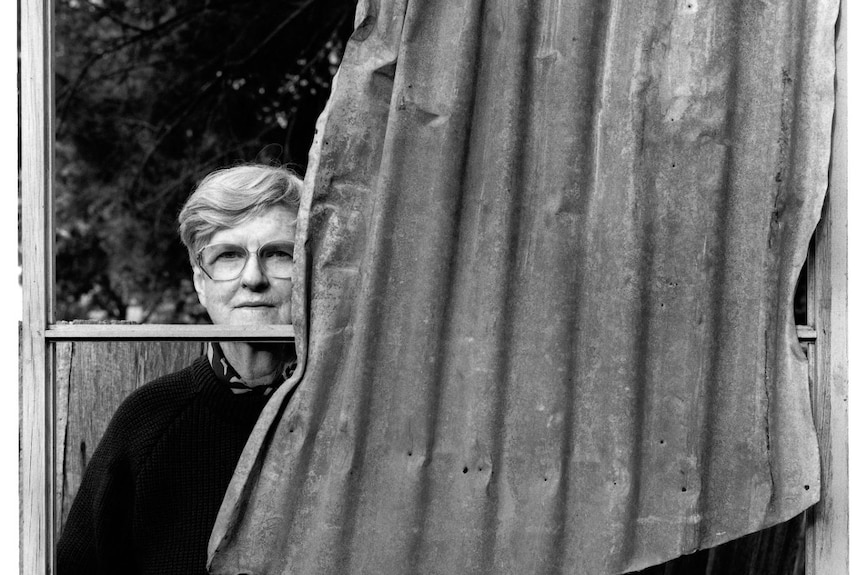 A black and white image of a woman with short grey hair standing helf-behind corrugated iron.
