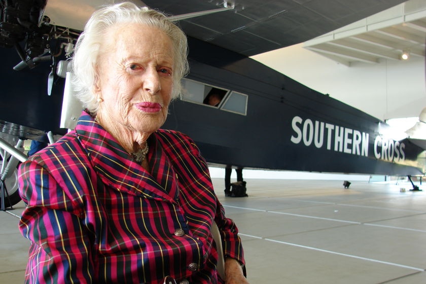 An elderly woman with white hair wears a striking blouse as she sits in a hangar near a vintage aircraft.