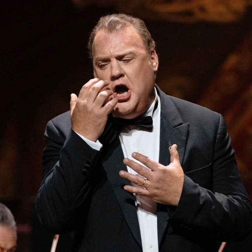 Sir Bryn Terfel performs onstage in a tailcoat, singing with orchestra musicians visible behind him.