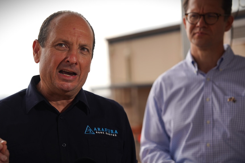 A man standing in a dark blue polo shirt left of frame mid-speech.