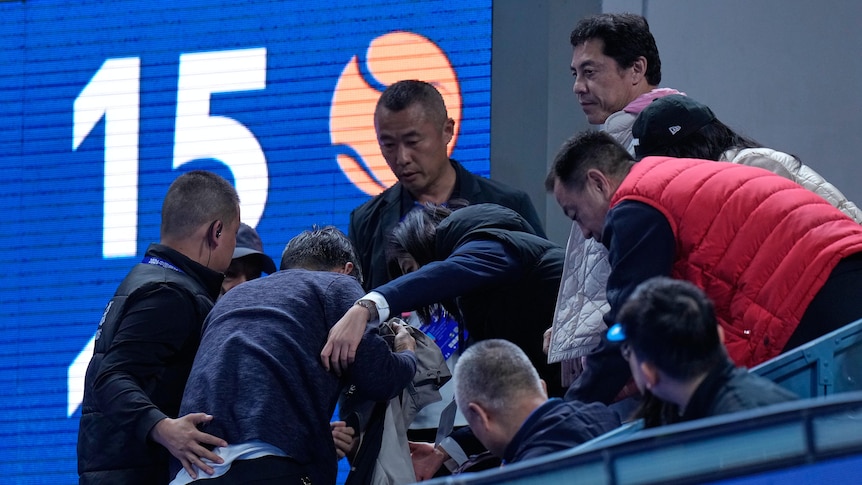 A group of people gather around an injured man (obscured) in the stands at a tennis tournament.