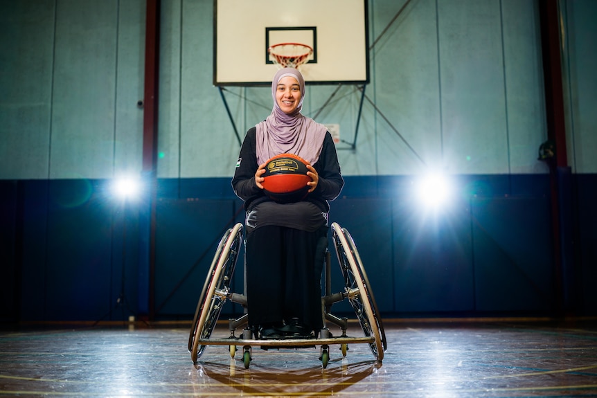 Reema El Qassem smiles as she sits in her wheelchair holding a basketball under a basketball hoop.