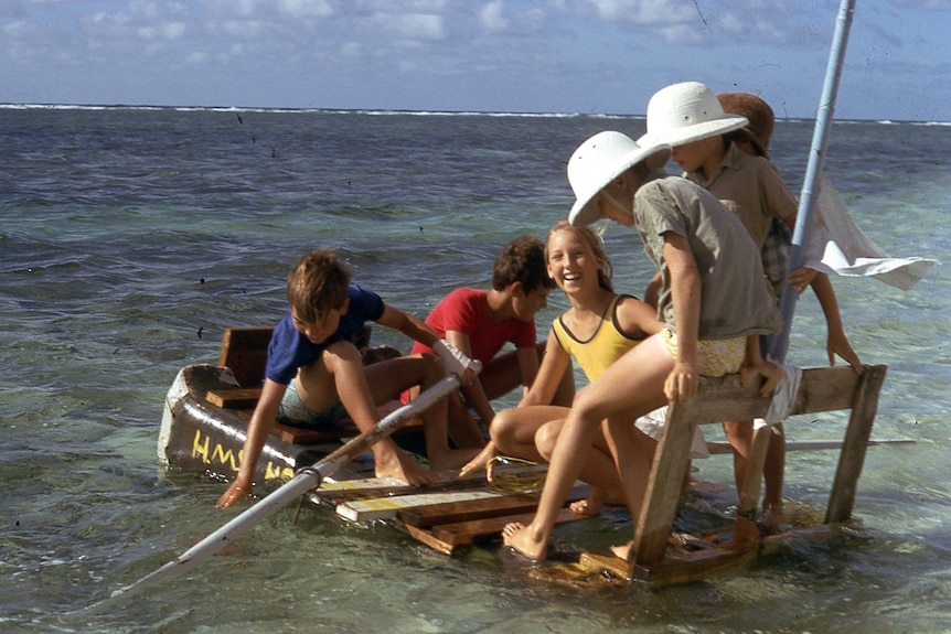 Five children on a homemade raft in shallow water.