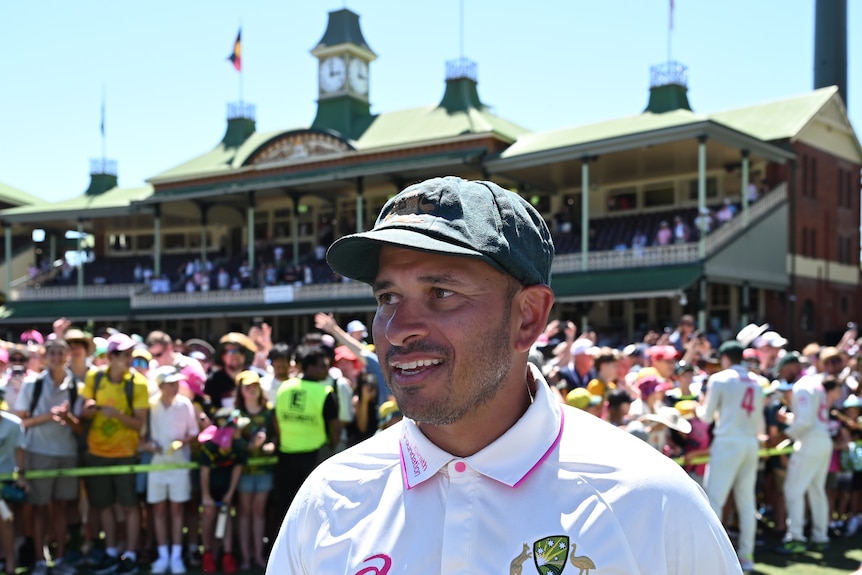 Usman Khawaja wears cricket whites with pink detailing as well as a baggy green cap. The crowd gathers behind in the stands.