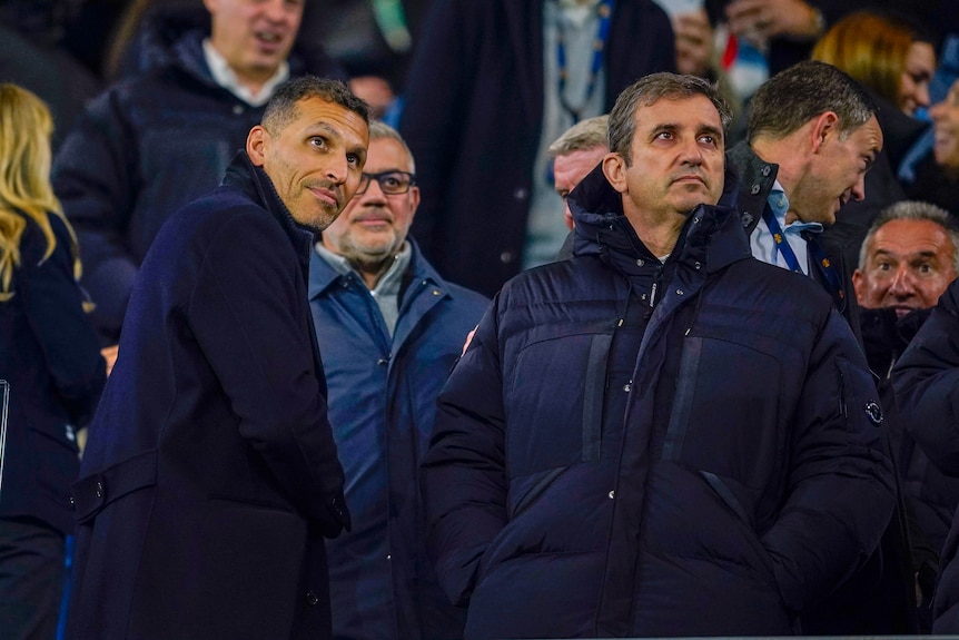 Two men in coats standing on the side of a football pitch before a match