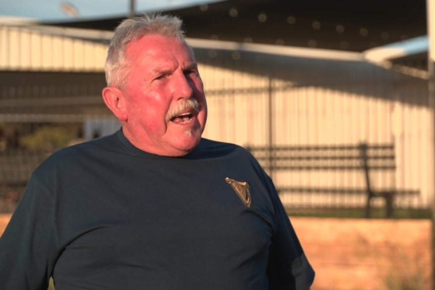 A man in a black tshirt is standing outside, in front of tin sheds.