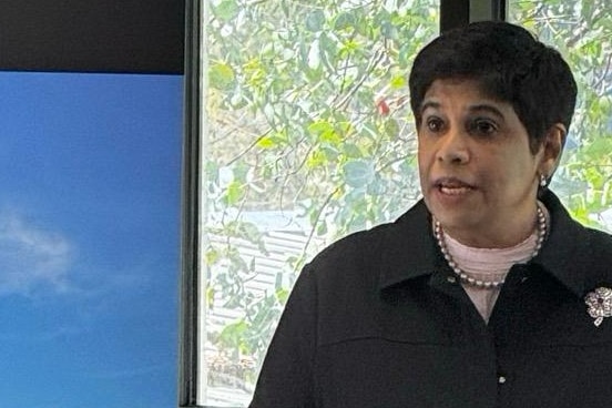 A woman with short black hair stands behind a lectern speaking to people out of frame