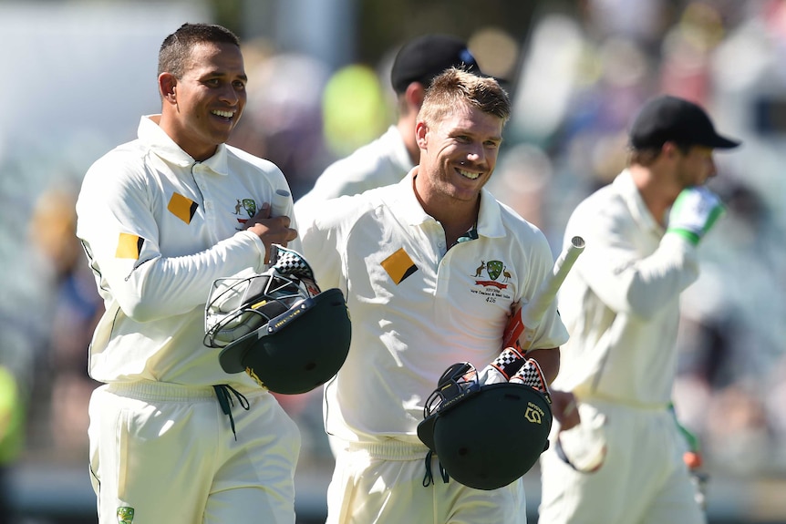 Dave Warner smiles big wearing reflective sunglasses with arm around Usman Khawaja. Both in cricket whites and green caps.