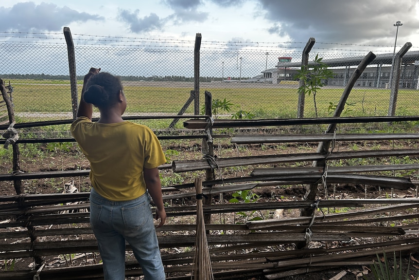 A young woman stands behind a fence with an airport on the other side. 