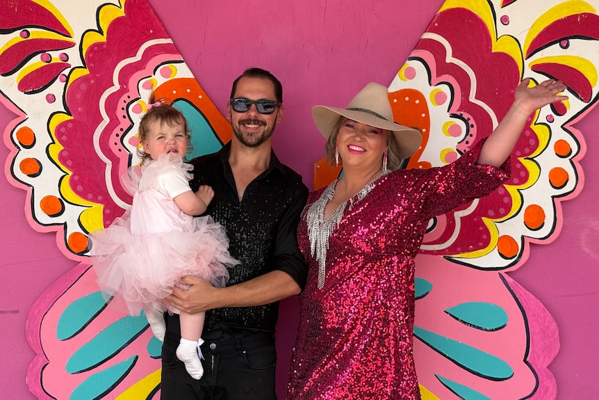 Kelly wears a bright pink sequined blazer and poses with her partner and child in front of a butterfly mural