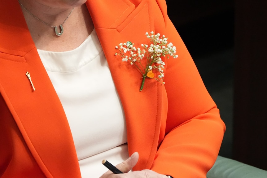 A close up of a small sprig of white flowers on lapel of a woman's orange blazer.