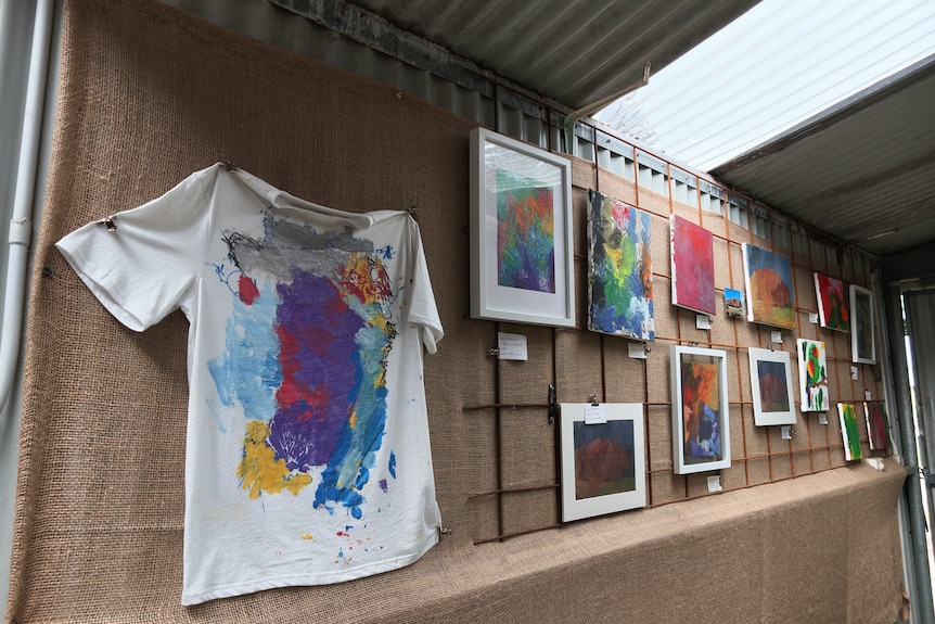 A hessian backdrop on a wall featuring a brightly painted T-shirt and artworks.