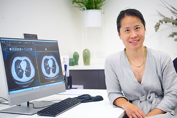 Alison Zhang, wearing a grey dress, is next to a computer and smiles at the camera.