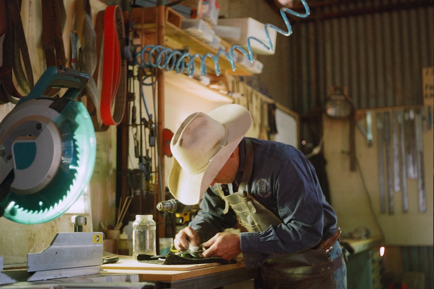 A man in a cowboy hat polishes a knife in a shed