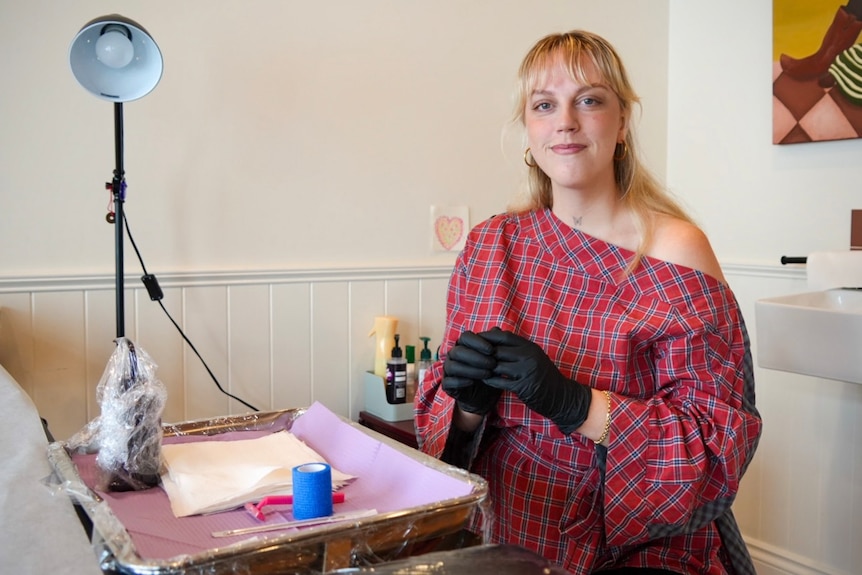 Blonde woman wearing a red checkered top with tattoo tray set up in front of her, putting on gloves and smiling at camera