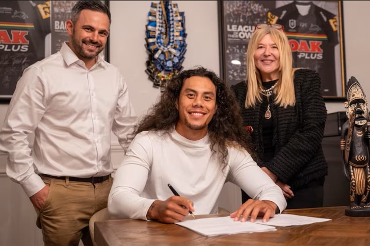 A young Samoan man wearing a white turtle neck with his hair out holds a pen over paper on a table with two people standing.