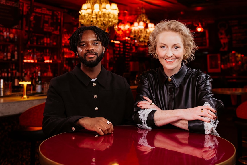Ghanian-Australian musician Genesis Owusu and Zan Rowe sit posing at a table at a bar table under chandeliers