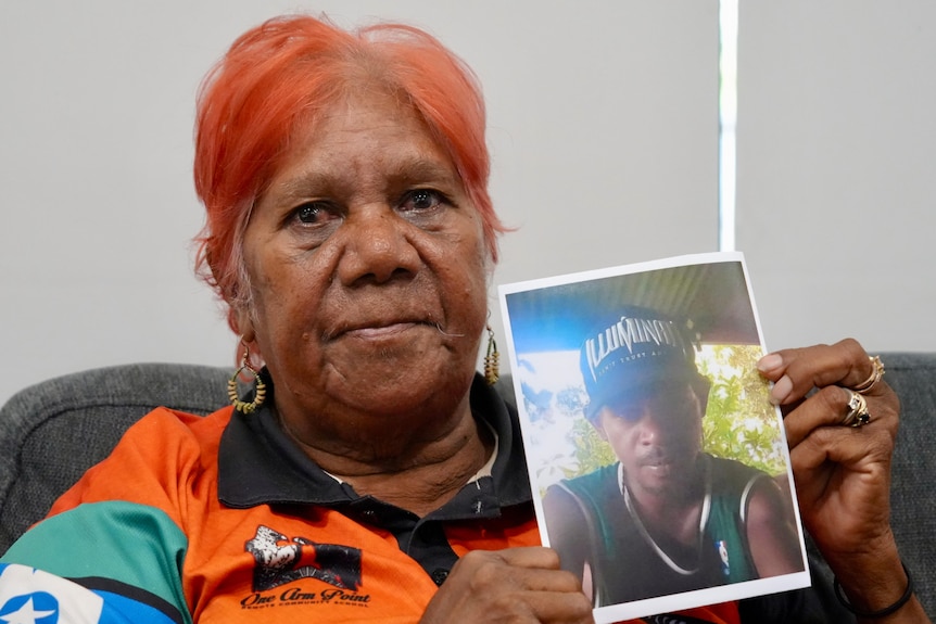 A woman with short hair holds a photo of a young man while looking sad.