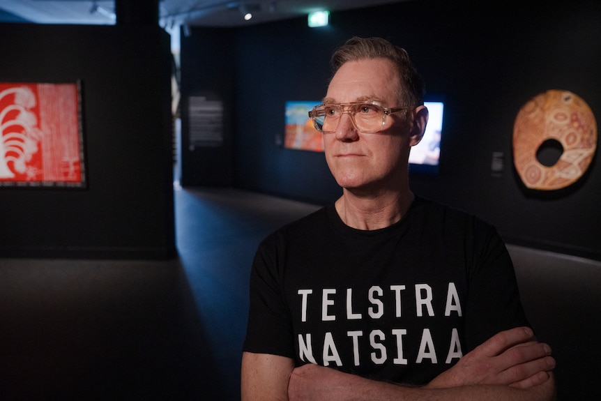 A man in an art gallery wearing glasses and a shirt that reads NATSIAA, he has a neutral facial expression.