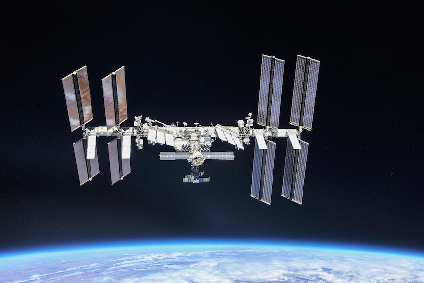A space craft with a central hub and broad rectangular wings sits floating above Earth