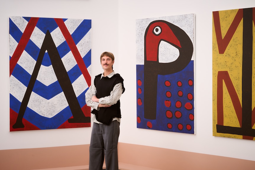 A person with a vest and moustache stands in a gallery room in front of large colourful graphic artwork