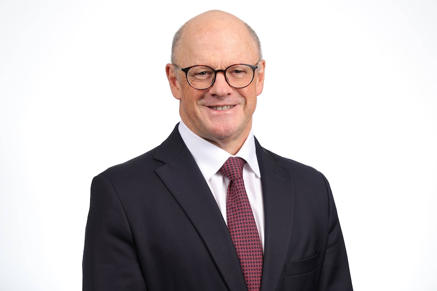 A man in a suit stands smiling in front of a white background.