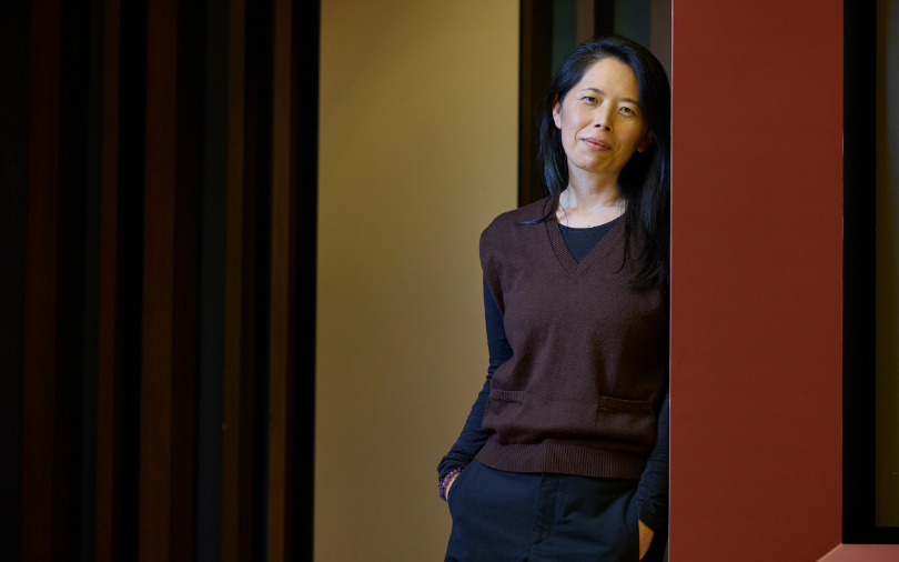 A woman with long dark hair stands in a doorway, wearing a brown V-neck sweater over a black shirt and dark pants. She has a calm, slight smile and looks directly at the camera. The background features vertical wooden slats and warm-toned walls.