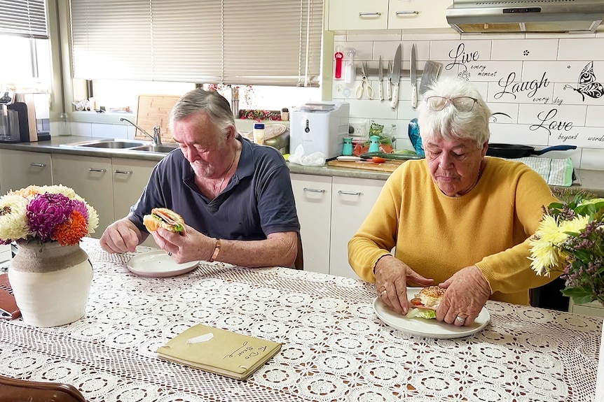 A couple sit at their dining table eating sandwiches