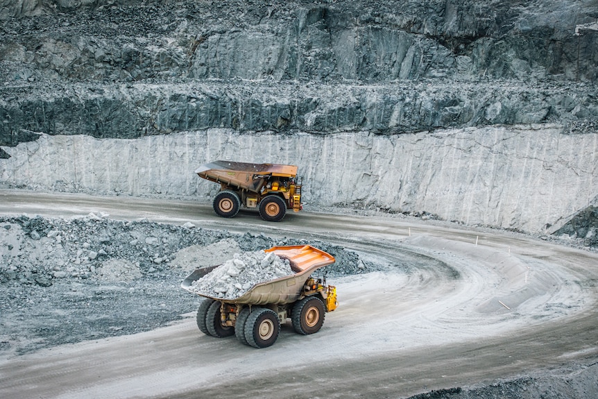 two massive dump trucks move along a dirt road that winds up an open pit mine