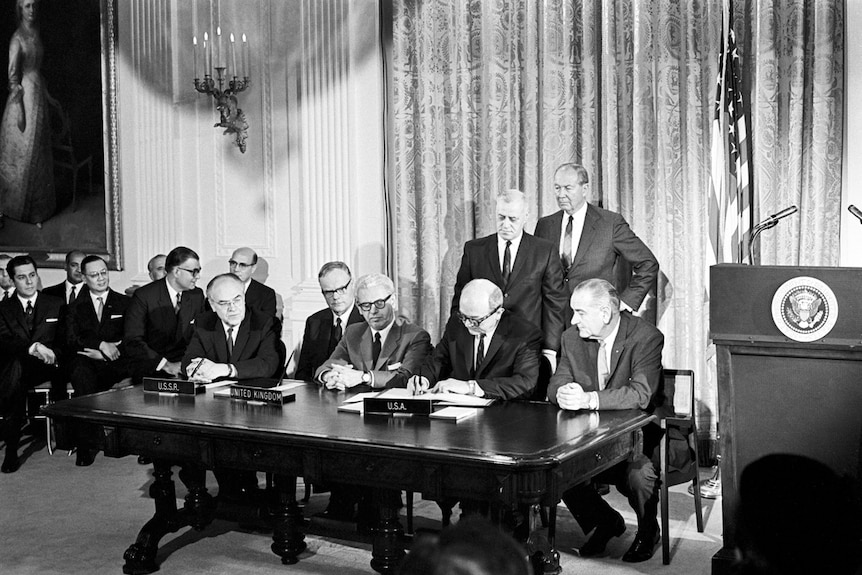 In a black and white photo a group of men sign a document.
