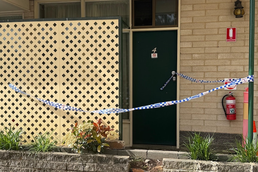 Police tape surrounds a motel room with a green door and cream brick.
