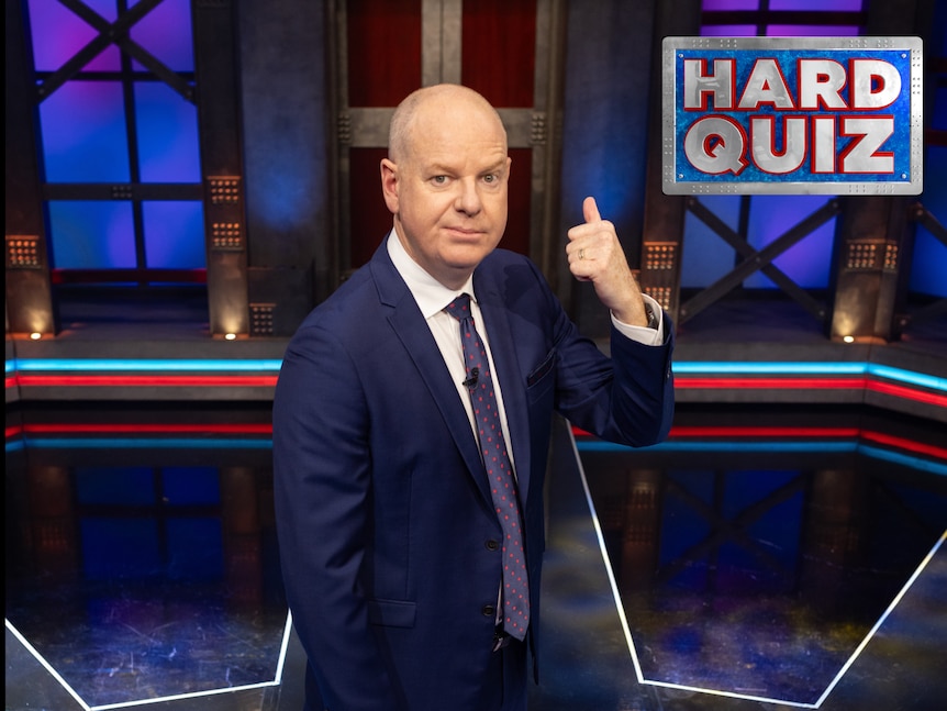 Tom Gleeson on the set of Hard Quiz gesturing behind him with a serious expression.