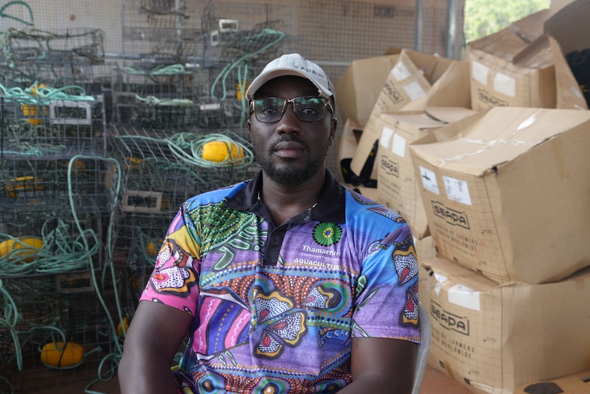 A Black man wearing a colourful shirt. There are several crab pots in the background and large cardboard boxes.