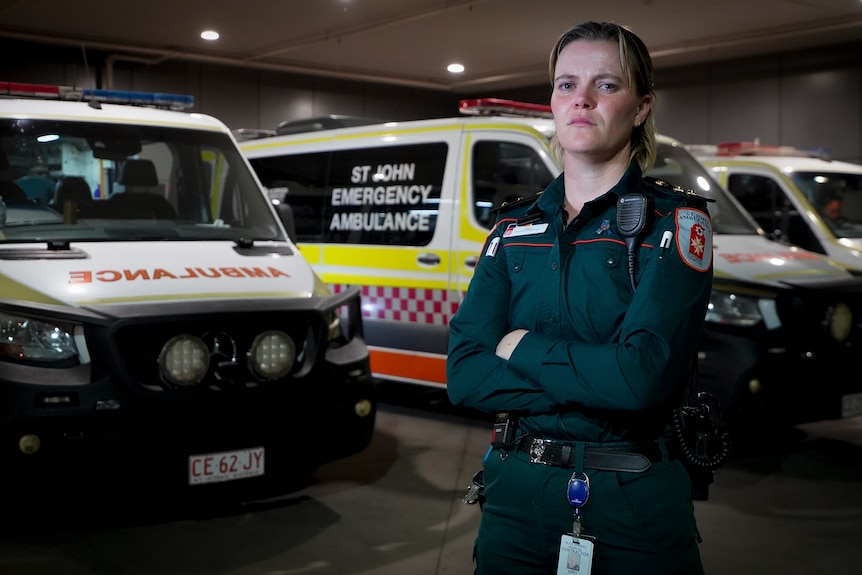 Abbey Studley wearing a green paramedic uniform stands in front of a row of ambulances with her arms folded