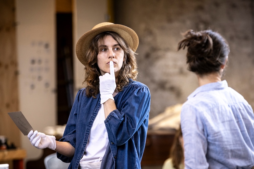 In a rehearsal room, Nathalie Morris, in a hat and white gloves, places one finger to her lips in thought.