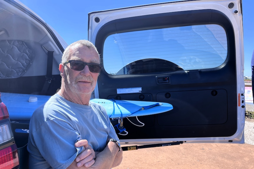 A man sits with crossed arms in the back of a car, a surfboard next to him