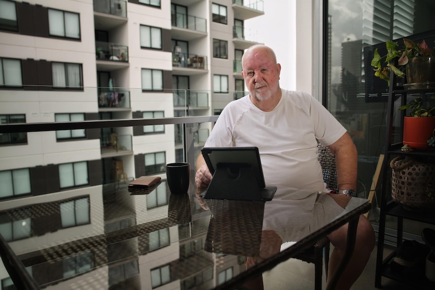 A man sitting at a table on a balcony with an apartment building visible behind him.