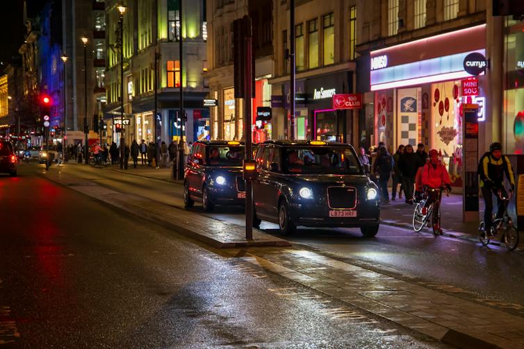 Cars driving on the street, people walking on the sidewalk with restaurants, retail stores and bars at night on the westside in London, England.