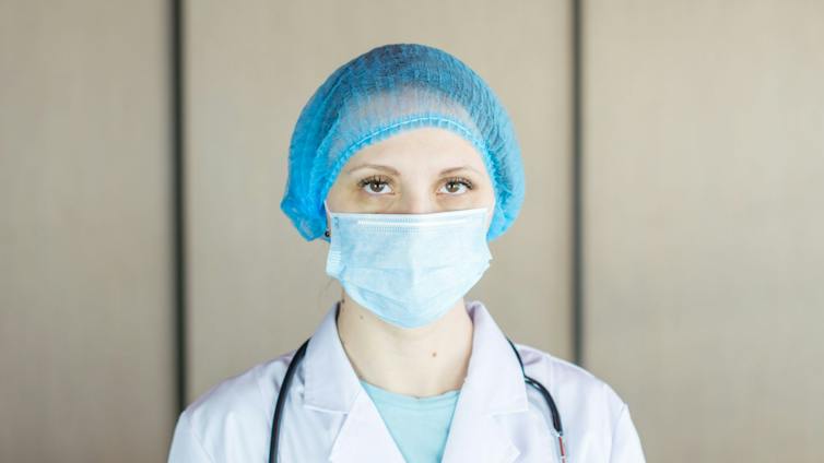 A woman wearing a white coat and blue PPE head covering and face mask, with a stethoscope around her neck