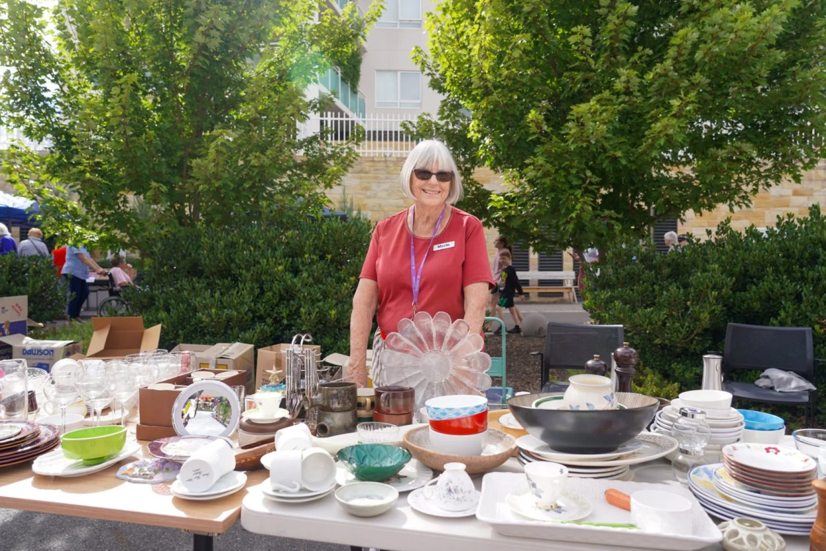 a person wearing a red top and sunglasses standing at a market stall of dishes on a table outside